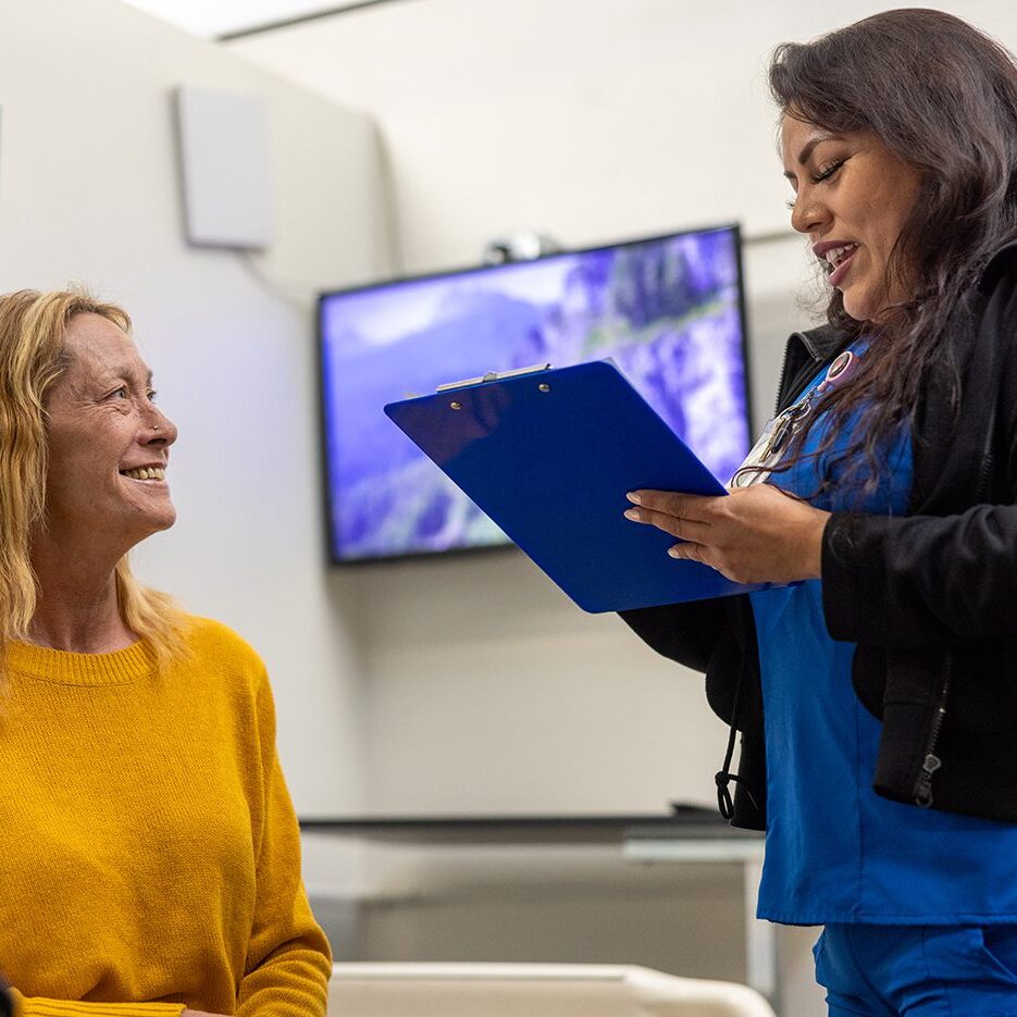 A healthcare professional stands holding a blue clipboard, smiling and talking with a seated woman in a yellow sweater in a clinical setting.