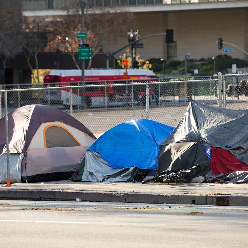 Several tents and makeshift shelters are set up on a sidewalk in an urban area, with a chain-link fence and city buildings in the background.