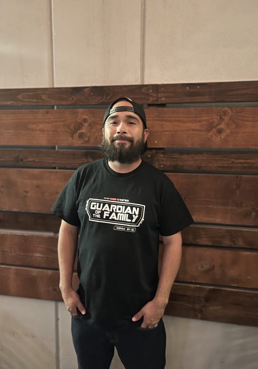 A man with a beard wearing a black Guardian of the Family t-shirt and a backward cap stands in front of a wooden wall, looking at the camera.