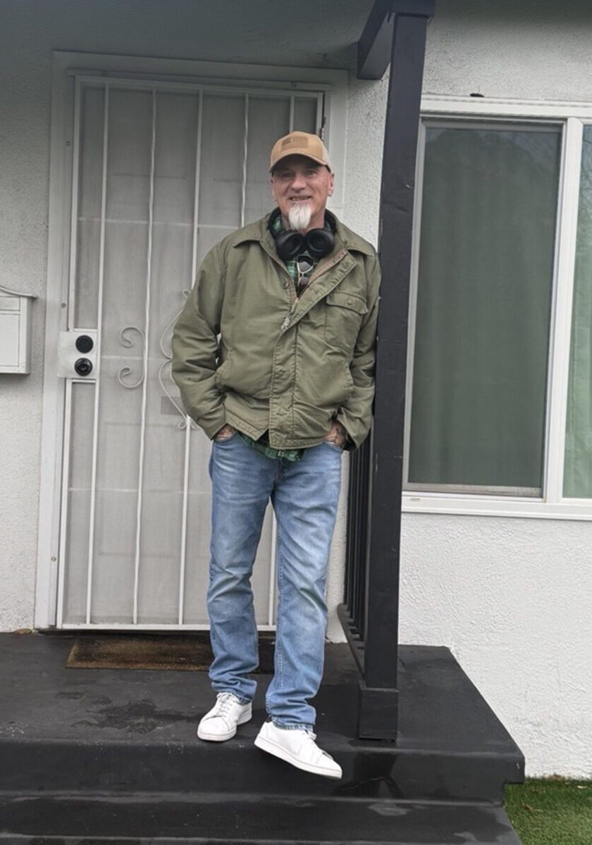 A man in a green jacket, blue jeans, and white sneakers stands on a black porch in front of a white door, with headphones around his neck and a tan cap on his head.