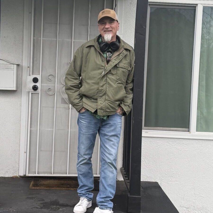 A man in a green jacket, blue jeans, and white sneakers stands on a black porch in front of a white door, with headphones around his neck and a tan cap on his head.